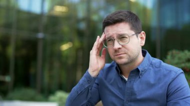Modern ofis binasının yakınındaki sokakta baş ağrısı çeken bir iş adamı. Üzgün yetişkin yakışıklı adam elleriyle başını tutuyor, şakaklarını ve alnını okşuyor ve masaj yapıyor. Kapat.