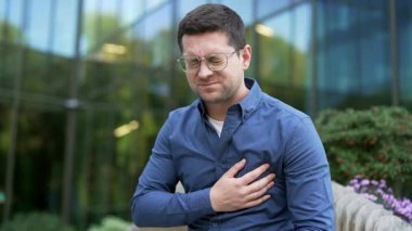 Hasta iş adamı kalp krizi geçiriyor, göğüs gerilmesinden muzdarip, ofis binasının yanındaki bankta oturuyor. Göğsünü elleriyle tutup masaj yapan üzgün bir adamın acil tıbbi müdahaleye ihtiyacı var. Kapat.