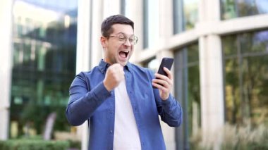 Mutlu iş adamı, bir iş yeri binasının yanında cep telefonundan harika haberler okuyarak başarıyı kutluyor. Gülümseyen adam neşeli bir mesaj aldı, zafer işareti yaptı, evet.