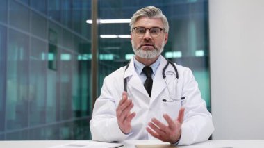 Webcam görüntüsü. Yetişkin bir doktor hastane ofisinde kameraya bakarak konuşuyor. Palto giyen tıp doktoru konferansta ya da hasta ile online istişarede uzaktan iletişim kurar