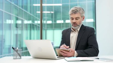 Bilgisayarla video konferansı seyreden odaklanmış işadamı iş yerindeki iş yerindeki not defterine notlar alıyor. Resmi kıyafetli olgun girişimci uzaktan eğitimleri dinliyor.