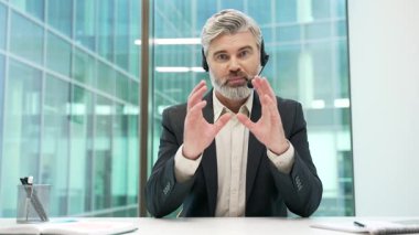 Webcam görüntüsü. Kulaklık takmış kendine güvenen bir iş adamı ofiste kameraya bakarak video görüşmesi yapıyor. Olgun girişimcilerin iş görüşmesi olur. Takım elbiseli müdür çevrimiçi konferansta uzaktan konuşuyor.