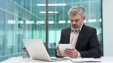 İşadamı, iş yerindeki masasında dijital tablet kullanıyor. Resmi takım elbiseli girişimci başvuruda çalışır, dizüstü bilgisayardaki verileri kontrol eder, bir görev veya projeyle ilgilenir