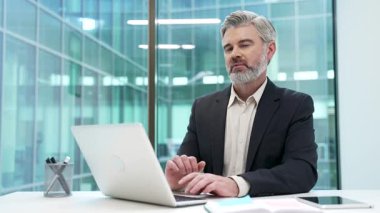 İş yerindeki çalışma masasında resmi takım elbiseli gülümseyen olgun iş adamının portresi. Mutlu, mutlu, sakallı, gri saçlı menajer dizüstü bilgisayarda çalışıyor ve kameraya bakıyor.