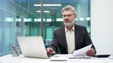 Modern iş ofisindeki çalışma masasında resmi takım elbiseli gülümseyen olgun iş adamının portresi. Başarılı, kendine güveni tam bir finansör. Bilgisayarda çalışıyor ve kameraya bakıyor.