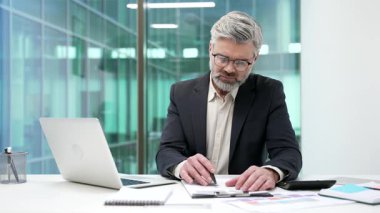 İş yerindeki bir masada oturan kendine güvenen işadamı belgeleri imzalıyor. Resmi takım elbiseli yetişkin patron, klasördeki belgelere bakar, kontrol eder ve kalemle sözleşme imzalar.