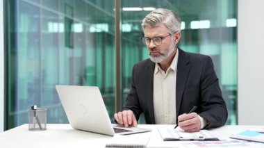 Yetişkin işadamı, iş yerindeki çalışma masasında oturan dizüstü bilgisayarını kullanarak belgeleri dolguluyor. Finansör evrak işleri, mali rapor, muhasebe ile meşgul.