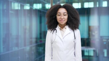 Hastane kliniğinde kameraya bakan beyaz önlüklü, gülümseyen Afrikalı Amerikalı kadın doktor portresi. Mutlu siyahi kadın doktor modern ofiste çapraz duruşlar sergiliyor.