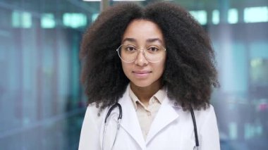 Hastane kliniğinde kameraya bakan beyaz önlüklü, gözlüklü, ciddi bir Afrikalı Amerikalı kadın doktorun portresi. Kendine güveni tam bir doktor ofisinde poz veriyor. Kapat.