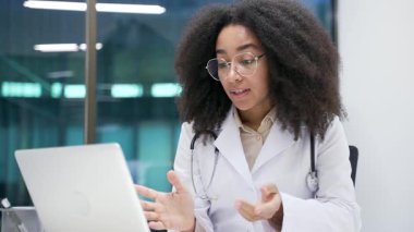 Kendine güvenen Afro-Amerikan kadın doktor hastane kliniğinde dizüstü bilgisayarla konuşuyor. Genç tıp çalışanı doktor, ofisinde oturan hastayla uzaktan görüşüyor. Kapat.