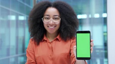 Mutlu Afro-Amerikan iş kadını iş yerinde yeşil ekranlı cep telefonu gösteriyor. Renk anahtarı, reklam için dikey şablon düzeni. Kameraya bakan kadın akıllı telefonu tutuyor. Yaklaş.