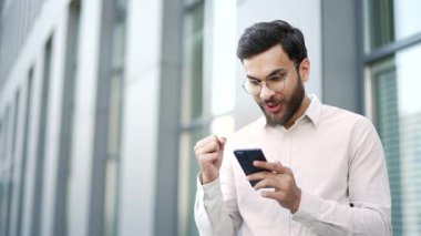 Memnun iş adamı ofis binasının yanındaki sokakta beklerken cep telefonundan harika haberler aldı. Gülümseyen yakışıklı girişimci akıllı telefondan olumlu bir mesaj okur. Kapat.