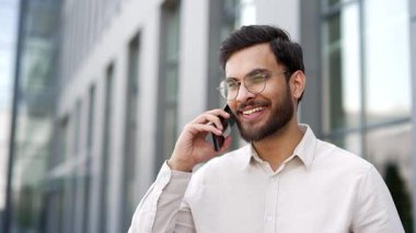 Ofis binasının yanındaki sokakta cep telefonuyla konuşan gülümseyen bir işadamı. Yakışıklı erkek, iş ortağı müşterisiyle iletişim kuruyor. Profesyonel adam akıllı telefondan konuşur. Kapat.