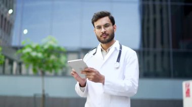 Beyaz önlüklü gülümseyen doktorun portresi hastane binasının yakınındaki kameraya bakarak dijital tablet kullanıyor. Gözlüklü, caddede poz veren yakışıklı, sakallı, erkek doktor.