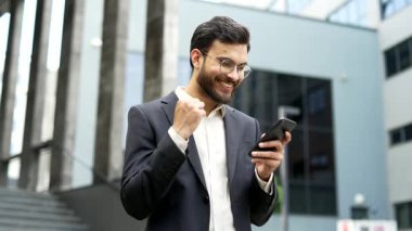 Memnun iş adamı ofis binasının yanındaki sokakta beklerken cep telefonundan harika haberler aldı. Resmi takım elbiseli mutlu bir girişimci akıllı telefondan olumlu bir mesaj okur.
