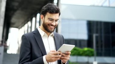 Gülümseyen işadamı ofis binasının yanındaki sokakta dikilirken dijital tablet kullanıyor. Resmi bir takım elbise içinde kendinden emin bir girişimci e-postaları kontrol ediyor, internette sohbet ediyor, mesajlaşıyor, internette geziniyor.