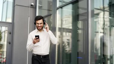 Kablosuz kulaklık takan mutlu işadamı, ofis binasının yakınındaki sokakta yürürken cep telefonundan müziğin keyfini çıkarıyor. Neşeli gülümseyen adam şarkı listesini açıyor ve şarkı dinliyor