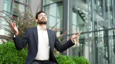 Takım elbiseli bir iş adamı, kapalı gözlerle meditasyon yapıyor. İş yerinin yanındaki bankta oturuyor. Mutlu erkek yoga yapıyor, derin nefes alıyor. İnsan stresten kurtulur, huzuru hisseder.