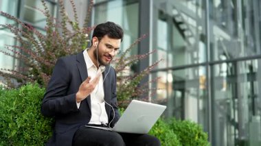 Resmi takım elbiseli bir iş adamı ofis binasının yanındaki bankta otururken bilgisayarla konuşuyor. Kulaklık takan yakışıklı girişimci müşteri veya iş ortağıyla çevrimiçi toplantı yapıyor.