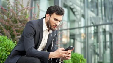 Odaklanmış işadamı, ofis binasının yanındaki bankta oturan cep telefonunu kullanıyor. Resmi takım elbiseli ve kulaklıklı kendine güvenen girişimci video veya yayın izliyor, konferansı dinliyor.