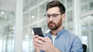 Memnun iş adamı iş yerinde otururken cep telefonundan harika haberler aldı. Gülümseyen yakışıklı girişimci akıllı telefondan olumlu bir mesaj okur. Kapat.