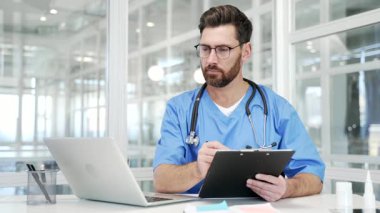 İşyerinde çalışan mavi ceketli bir doktor. Hastane masasında dizüstü bilgisayarını kullanarak veri yazıyor. Evrak işleri yapan doktor, form dolduran ya da reçete yazan.