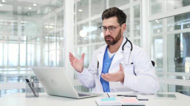 Hastane ofisinde dizüstü bilgisayarla konuşan beyaz önlüklü kendine güvenen bir doktor. Erkek doktor konferansta uzaktan iletişim kurar ya da hastayla çevrimiçi danışmanlık yapar