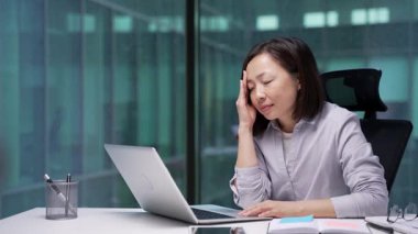 İş yerindeki çalışma masasında dizüstü bilgisayarla çalışırken baş ağrısı çeken Asyalı yorgun bir iş kadını. Üzgün kadın çalışan acı hisseder, masaj yaparken eli şakaklarını tutar.
