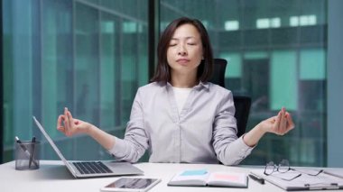 Asyalı iş kadını, modern iş ofisindeki işyerinde otururken gözleri kapalı meditasyon yapar. Bayan çalışanımız işe ara verdi. Sakin bir kadın dinlenir, rahatlar, stresten kurtulur, huzuru hisseder.