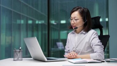 Kablosuz kulaklık takmış kendine güvenen Asyalı iş kadını ofiste oturan dizüstü bilgisayarla video konferansında konuşuyor. Kadın antrenör uzaktan konuşuyor, çevrimiçi toplantıda ya da antrenmanda iletişim kuruyor.