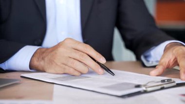 İş yerindeki bir masada bir erkeğin el imzalı belgelerini kapatıyoruz. Resmi takım elbiseli bir işadamı belgelerle dosyaya bakar ve sözleşmeyi kalemle imzalar.