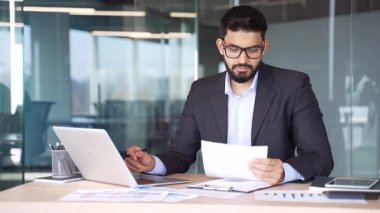 Resmi takım elbiseli meşgul işadamı iş yerinde otururken bilgisayar kullanarak evrak işleriyle uğraşıyor. Belgelerle çalışan düşünceli bir finansör, bir rapor hazırlıyor, muhasebe ile meşgul.