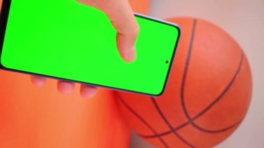 Turuncu basketbol topunun yanındaki yeşil ekranlı el kameralı akıllı telefonun dikey videosu