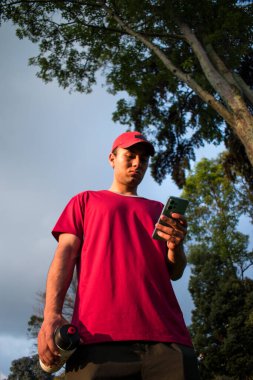 Kırmızı şapkalı ve elinde şişe olan bir adam gün batımında parkta mavi telefonuna bakıyor.