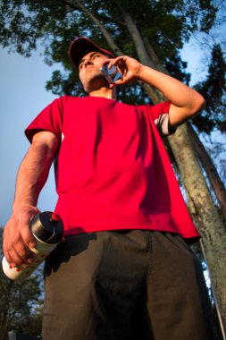 Kırmızı şapkalı ve elinde şişe olan bir adam gün batımında parkta mavi telefonuyla konuşuyor.