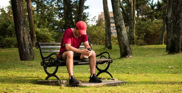 Man wearing red cap, taking rest  and holding a bottle in park bench at sunset