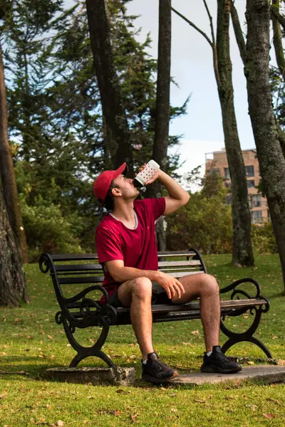 Man wearing red cap and drinking water from bottle in park bench at sunset