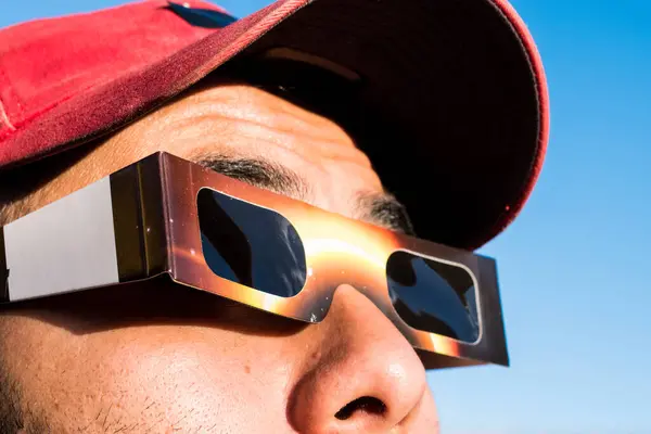 Şapka takan ve güneş tutulması gözlüğü takan insanın portresi, arka planda mavi gökyüzü olan güneşe bakıyor. 