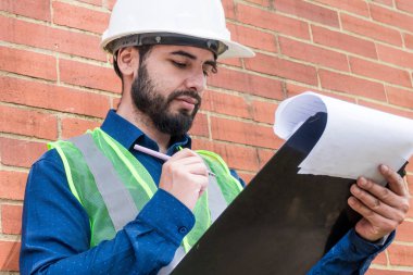 Latin inşaat işçisi, güneşli bir günde arka planda tuğla duvarlı belgeler arıyor.