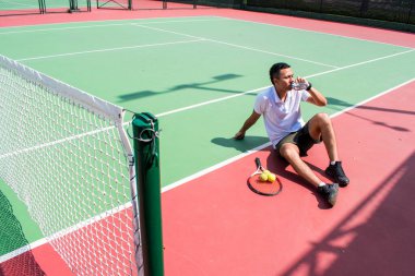 Genç latn tenisçisi güneşli yaz gününde tenis kortunda dinleniyor ve su içiyor.