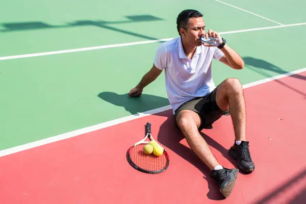 Young latn man tennis player resting and drinking water on tennis court in sunny hot day at summer