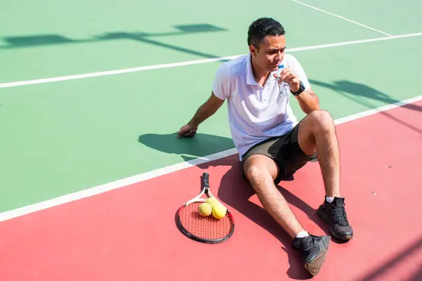 Young latn man tennis player resting and drinking water on tennis court in sunny hot day at summer