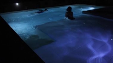 Havuzda iki kadın, gece mavi ışıkla aydınlatılıyor. 