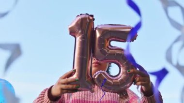 Partide 15 yıllık balonlar tutan bir kız. Arka planda mavi gökyüzü var.