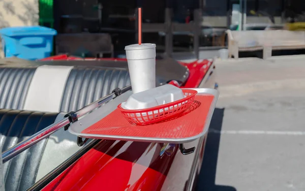 food tray on an antique red convertible vintage car with a styrofoam drink cup red straw and styrofoam hotdog holder. Selective focus on food tray and contents.