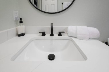 Lavabo tezgahında bir sabun şişesi, düzgünce katlanmış bir havlu ve arka plandaki sahneyi yansıtan bir ayna var..