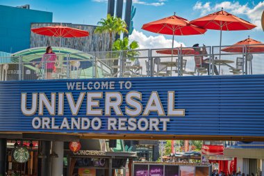 Orlando Florida Birleşik Devletleri 13 Mayıs 2025 Universal Orlando Tatil Köyü tabelasının üzerinde bir erkek telefonuna dolanmış turuncu şemsiyesinin altında bir kadın yakınında dururken oturuyor. Sadece editör kullanımı