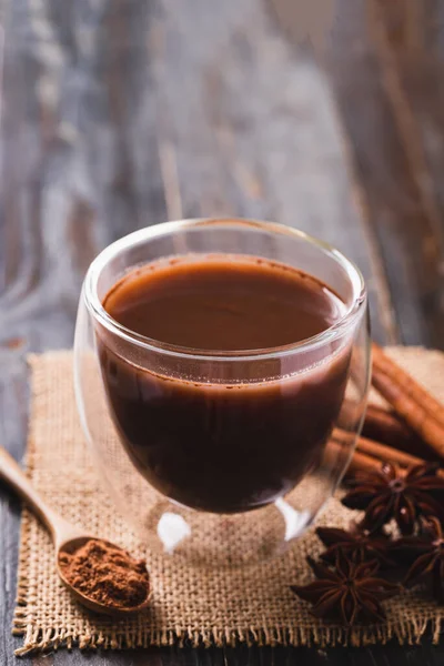 Hot cocoa in cup glass with cinnamon stick, star anise and cocoa powder on wooden background, Hot drink in winter season