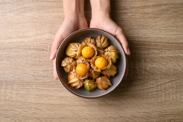 Cape gooseberry or golden berry (Physalis peruviana) in bowl with hand on wooden background, Table top view
