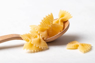 Tahta bir kaşığın üzerine çiğ farfalle makarna. Beyaz bir arka plana yerleştirilmiş papyon şeklinde makarna. İtalyan mutfak malzemelerini vurgulayan makarna.